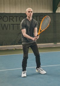 Grauem, kurzärmeligem Sportshirt und dunklen Hosen, getragen von einer Person, die einen orangefarbenen Tennisschläger auf einem blauen Hallenplatz hält.