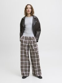 Braune Kunstlederjacke, graues enges T-Shirt und weit geschnittene karierte Hose in Weiß- und Brauntönen. Schwarze klobige Schuhe runden das Outfit ab.