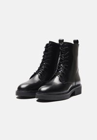 Botas de cuero negro hasta el tobillo con punteras redondas, cordones prominentes, suelas de goma planas y tiradores en la parte superior para ponérselas fácilmente. Textura suave y diseño elegante.