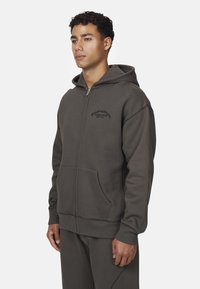 Felpa con zip grigia realizzata in tessuto felpato, dotata di tasca a marsupio, branding minimale e cuciture a contrasto lungo le cuciture.