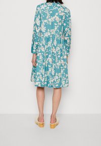 Turkosblommig klänning med långa ärmar, samlad midja och avslappnad passform. Har ett lätt, texturerat tyg och träklackar.