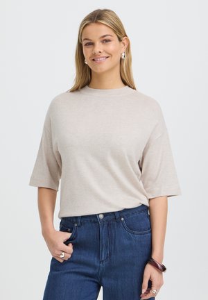 Žena s dlhými blond vlasmi sa usmieva, má na sebe béžový top s krátkym rukávom, modré džínsy, kruhové náušnice a náramok, stojí pred jednoduchým pozadím.