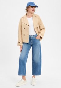 Veste beige à double boutonnage, t-shirt blanc, jeans bleu à jambes larges et baskets blanches. Casquette bleue avec logo. Fond uni.