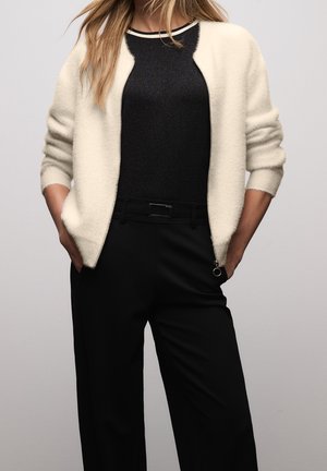 Vrouw draagt een witte, pluizige cardigan met ritssluiting over een zwarte top met witte bies en zwarte broek met hoge taille, handen in de zakken.