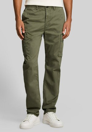 Mann trägt olivgrüne Cargohosen mit Seitentaschen und weißen Sneakers und steht vor einem einfarbigen Hintergrund.