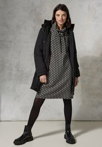 Schwarze Parka mit abnehmbarer Kapuze, getragen über einem schwarz-weißen Hahnentrittkleid. Schwarze Strumpfhosen und klobige schwarze Stiefeletten.