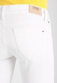 Bílé džínové kalhoty s polovičním pasem, opatřené zadními kapsami a béžovým koženým štítkem s nápisem "TOMMY JEANS" na pasu.