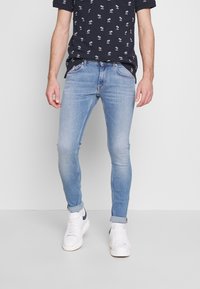 Ljusblå skinny jeans med en blekt textur och subtila sömnadsdetaljer, kombinerat med en svart t-shirt med kort ärm som har ett vitt palmträdsmönster.