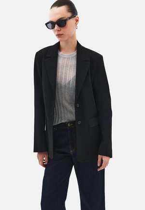 Sort blazer med hakket revers, to knapper og forlommer, båret over en sølvfarvet mesh-top og mørke jeans, suppleret med solbriller.
