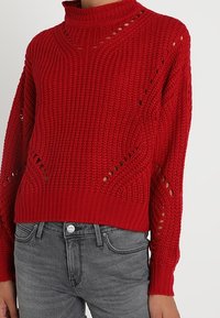 Pull en maille rouge avec un col montant, présentant des détails en point ajouré et des poignets côtelés. Coupe courte et ample, associée à un jean gris.
