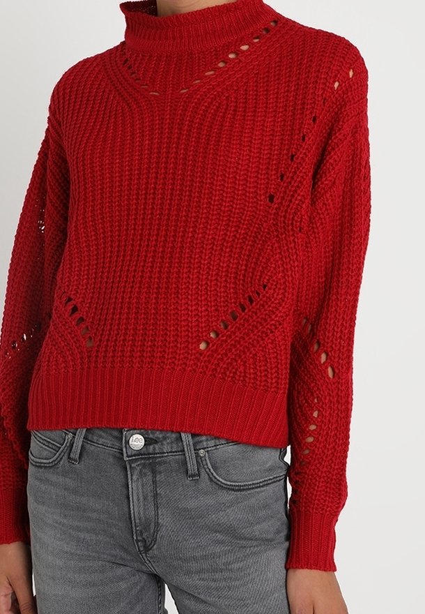 Pull en maille rouge avec un col montant, présentant des détails en point ajouré et des poignets côtelés. Coupe courte et ample, associée à un jean gris.