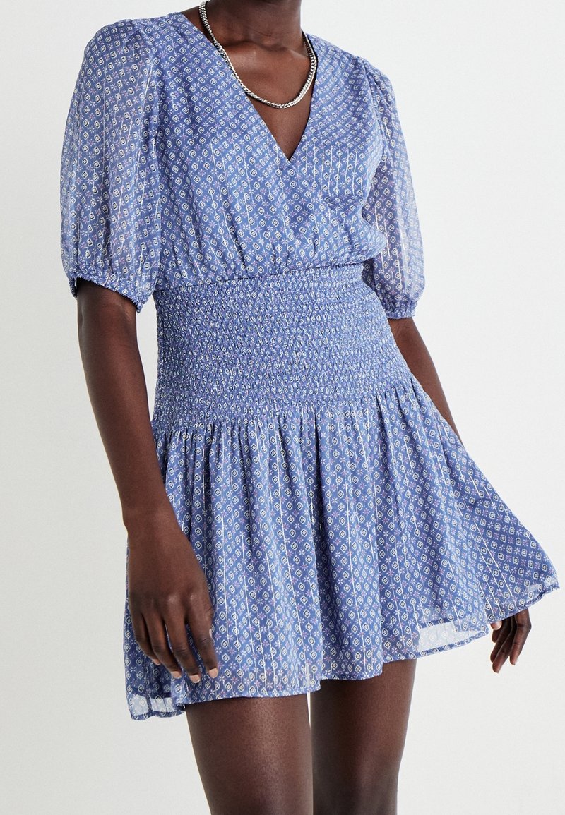 Femme portant une robe portefeuille bleue à motifs avec des manches bouffantes, une taille smockée et une jupe évasée, accessoirisée d'un collier chaîne en argent.