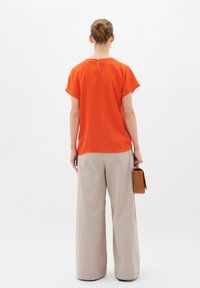 Top arancione a maniche corte, vista posteriore, con dettaglio a occhiello sul collo; abbinato a pantaloni beige a gamba larga e una borsa marrone.