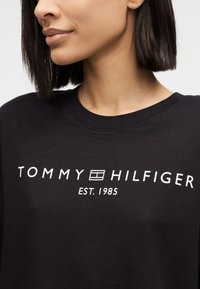 Γυναίκα με κοντά σκούρα μαλλιά, που φοράει μαύρο μπλουζάκι με λαιμόκοψη και λευκό κείμενο "Tommy Hilfiger Est. 1985" και λογότυπο στο στήθος.