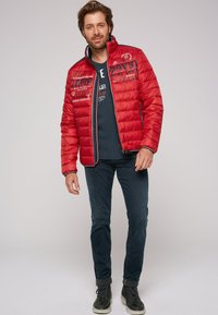 Giacca imbottita rossa con colletto alto, caratterizzata da grafiche testuali e piping contrastanti in navy. Indossata sopra una camicia blu scuro e abbinata a jeans scuri.