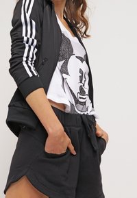 Svart zip-up bomberjacka med vita ränder, vit t-shirt med grafisk utskrift som föreställer en karaktär, och svarta shorts med dragsko och fickor.