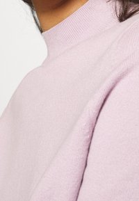 Pull rose clair avec un col haut et une texture côtelée, mettant en valeur un tissu doux et des lignes fluides. Gros plan sur le col.