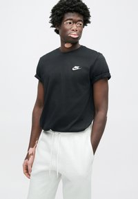 Juoda medvilninė marškinėliai su susuktomis rankovėmis ir baltu Nike logotipu ant krūtinės, derinama su šviesiai pilkomis sportinėmis kelnėmis. Tekstūruota medžiaga, laisvas modelis.