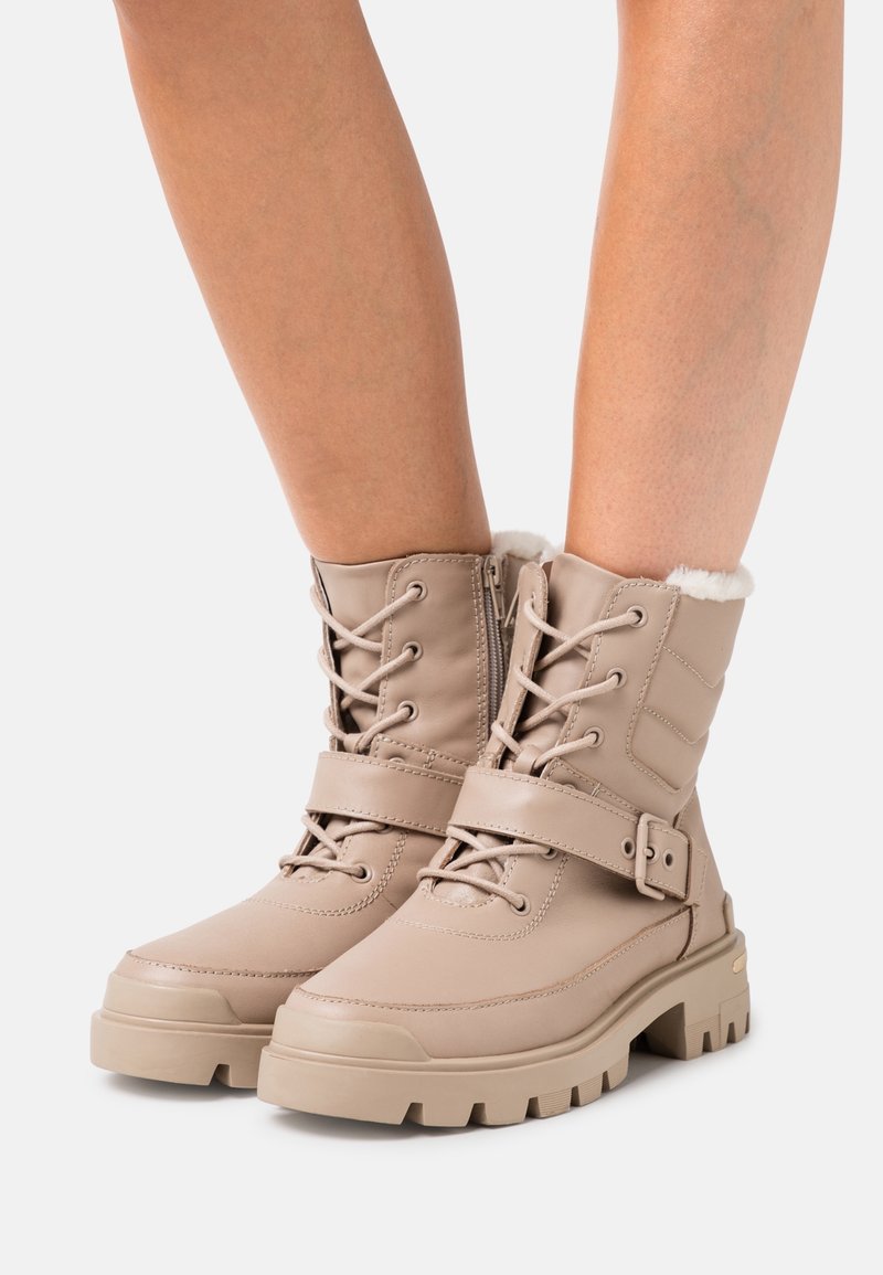 Beige winterboots met een zachte faux-fur voering, een vetersluiting aan de voorkant, een verstelbare band en een stevige rubberen zool met een geribbelde textuur.