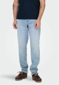 Man draagt lichtblauwe rechte jeans, donkerbruine leren bootsschoenen met roze veters en een donkerblauw shirt, staand tegen een witte achtergrond.