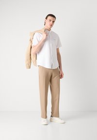 Camicia bianca a maniche corte con colletto a punta, pantaloni beige e sneakers avorio, con giacca beige sulle spalle. Design semplice e pulito.