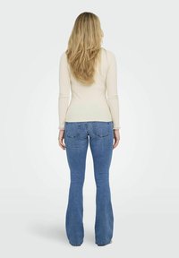 Ljust beige långärmad topp med volangförsedda ärmar, tillsammans med åtsittande blå flare jeans sedd bakifrån. Slät textur, avslappnad design.