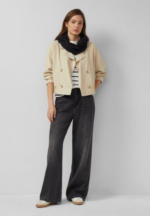 Frau trägt beige cropped Jacke, schwarzen Schal, gestreiftes Hemd, weit geschnittene schwarze Jeans und weiße Sneakers und steht vor einer schlichten hellgrauen Wand.