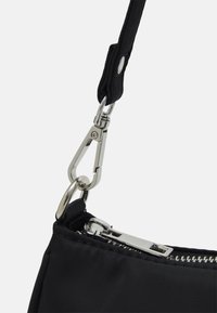 Černý nylonový popruh přes rameno připevněný stříbrným kovovým zapínáním, s zipem a prošívanou texturou podél okraje.