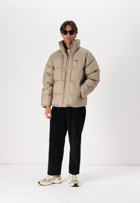 Giacca puffer beige con colletto alto e zip frontale, caratterizzata da un piccolo logo nero sul petto, abbinata a pantaloni di corduroy neri e sneakers beige.