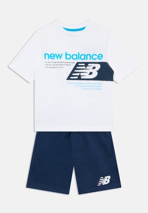 T-shirt blanc New Balance avec un logo bleu et une inscription bleu marine, assorti à un short bleu marine orné d’un logo blanc, sur un fond uni.