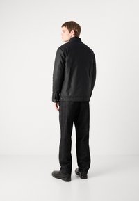 Schwarze isolierte Jacke mit hohem Kragen, strukturiertem Stoff und Taschen an den Seiten, getragen über einer dunklen, geraden Hose und schwarzen Stiefeln.