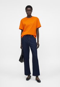 Γυναίκα φοράει φωτεινό πορτοκαλί oversized μπλουζάκι, σκούρα μπλε τζιν με φαρδιά πόδια, μαύρα μπαλαρίνια και κρατάει μια μαύρη τσάντα σε απλό ανοιχτό φόντο.