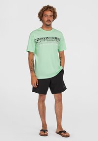 Camiseta de manga corta verde clara con un diseño de logo en blanco y negro, combinada con pantalones cortos negros y chanclas negras.