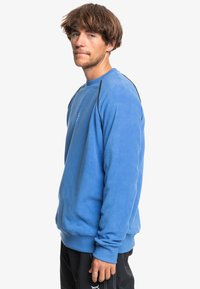 Sweatshirt en polaire bleu avec un col rond, des manches longues et des accents noirs sur les épaules, comportant un logo discret sur la poitrine.