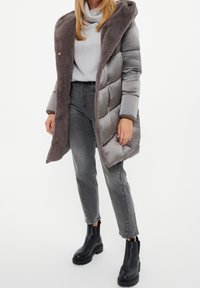 Abrigo acolchado gris con capucha de felpa, con acabado brillante y diseño acanalado. Combinado con jeans grises y botines negros.