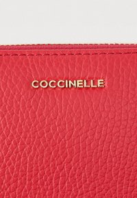 Rode textuur leren oppervlak met een gouden metalen rits en gouden logo letters die "COCCINELLE" spellen, gecentreerd onder de rits.