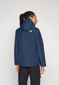 Veste imperméable bleu marine avec capuche et logo au dos. Elle présente des poignets élastiques et un ourlet droit. Texture lisse, design contemporain.