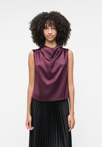 Top senza maniche di satin bordeaux con scollatura a cappuccio, abbinato a una gonna nera plissettata. Texture liscia, vestibilità rilassata, design minimalista.