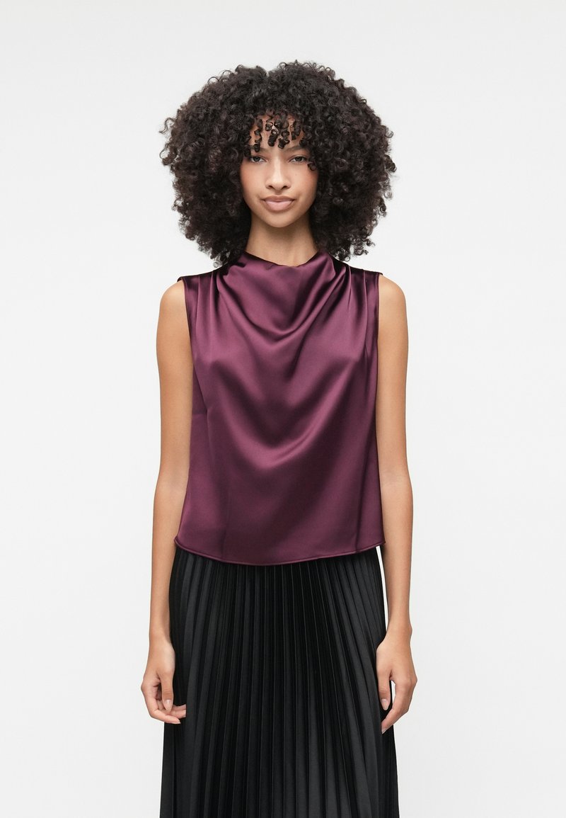 Top senza maniche di satin bordeaux con scollatura a cappuccio, abbinato a una gonna nera plissettata. Texture liscia, vestibilità rilassata, design minimalista.