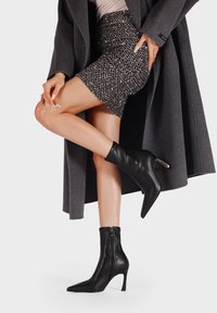 Stivali alla caviglia in pelle nera con punta affusolata e tacco a stiletto, caratterizzati da una cerniera laterale e una superficie liscia, abbinati a una gonna glitterata.