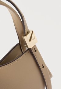 Μπεζ δερμάτινη τσάντα με σκούρα καφέ άκρες και χρυσό κούμπωμα χαραγμένο με τη φράση "kate spade new york" στο λουρί.