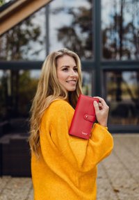 Red leather wallet with a button closure, smooth texture, and a logo on the front, held against a bright yellow sweater.