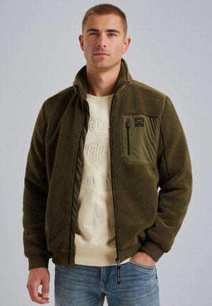 Donkergroene fleecejack met een ritsborstzak, geribbelde manchetten en een gladde kraag, over een lichtbeige sweatshirt gedragen, in combinatie met denim.