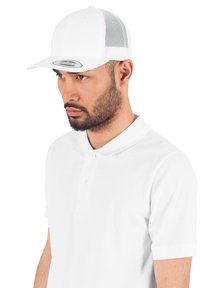 Gorra snapback blanca con lados de malla, visera curvada y un parche de marca ovalado y brillante en la parte frontal. Llevada con una camiseta polo blanca.