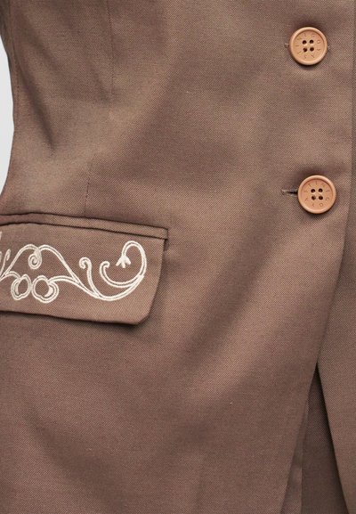 Καφέ ύφασμα με δύο ανοιχτό καφέ κουμπιά και τσέπη με καπάκι που φέρει άσπρο κεντητό σχέδιο στροβίλου σε ένδυμα.