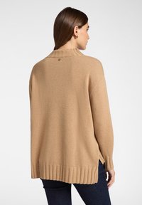 Chunky stickad tröja i beige, med ribbad krage och muddar, sidoinsnitt vid fållen och en liten logotypdetalj på baksidan.