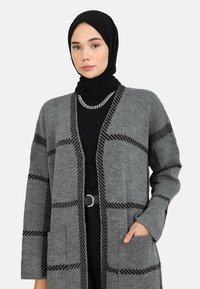 Cardigan grigio con motivo a strisce nere, aperto davanti, maniche lunghe e due tasche frontali, indossato sopra una maglia nera e accessorato con una catena d'argento.