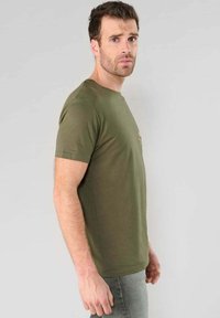 Hombre con cabello corto castaño y barba, vistiendo una camiseta verde oliva y jeans grises, de pie de perfil contra un fondo claro y liso.