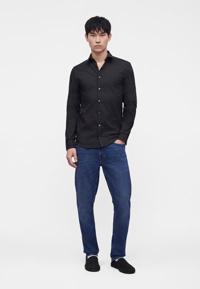 Mann steht mit der Hand in der Tasche, trägt ein schwarzes Hemd mit Knöpfen, blaue Jeans und schwarze Slipper vor einem schlichten weißen Hintergrund.