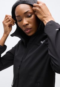 Μαύρο μπουφάν softshell με ψηλή γιακά και κουκούλα. Διαθέτει τσέπες με φερμουάρ και λεία, ματ υφή. Μινιμαλιστικός σχεδιασμός χωρίς εμφανή λογότυπα.